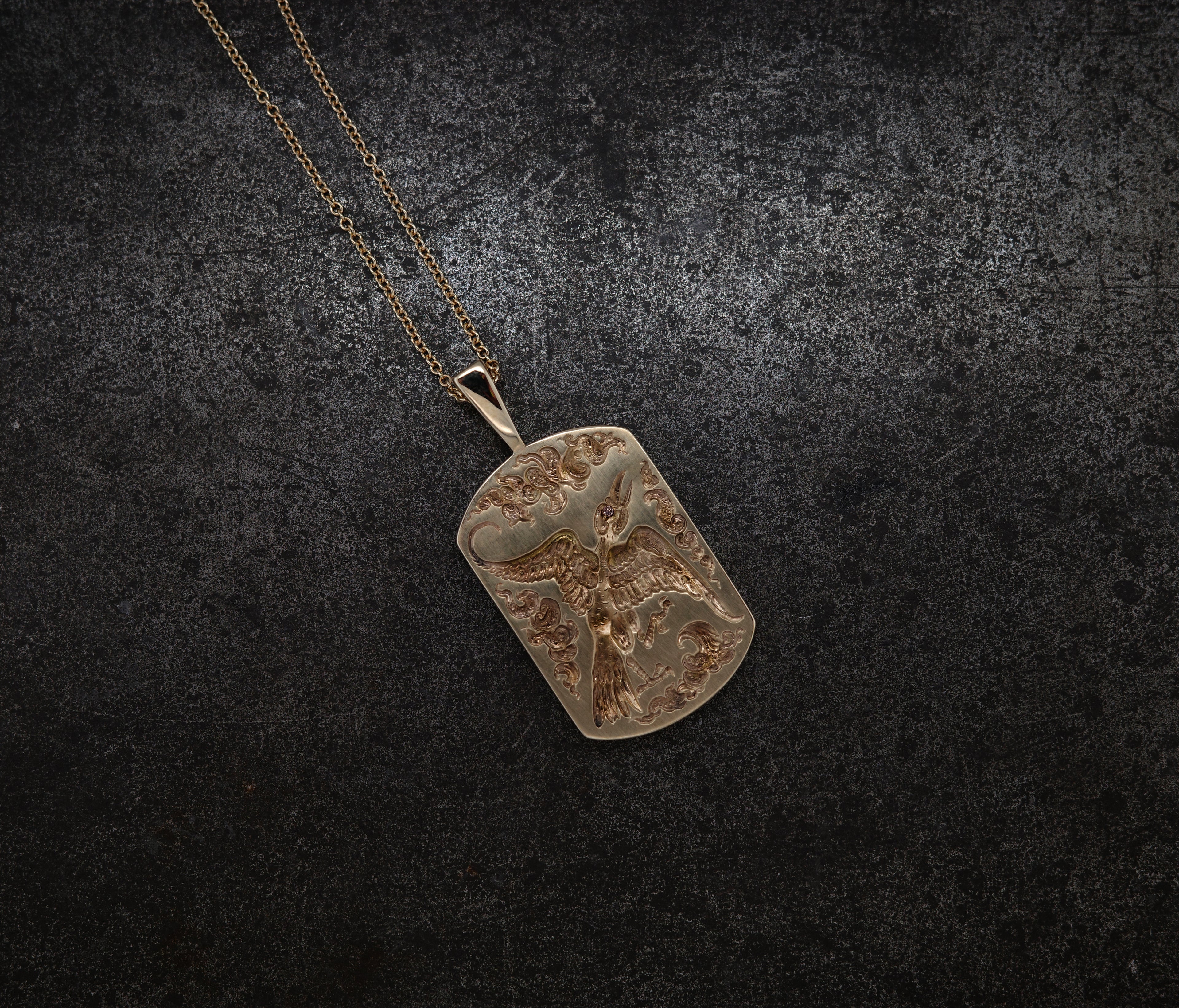 Gold bird pendant with intricate engravings hanging from a delicate gold chain on dark background