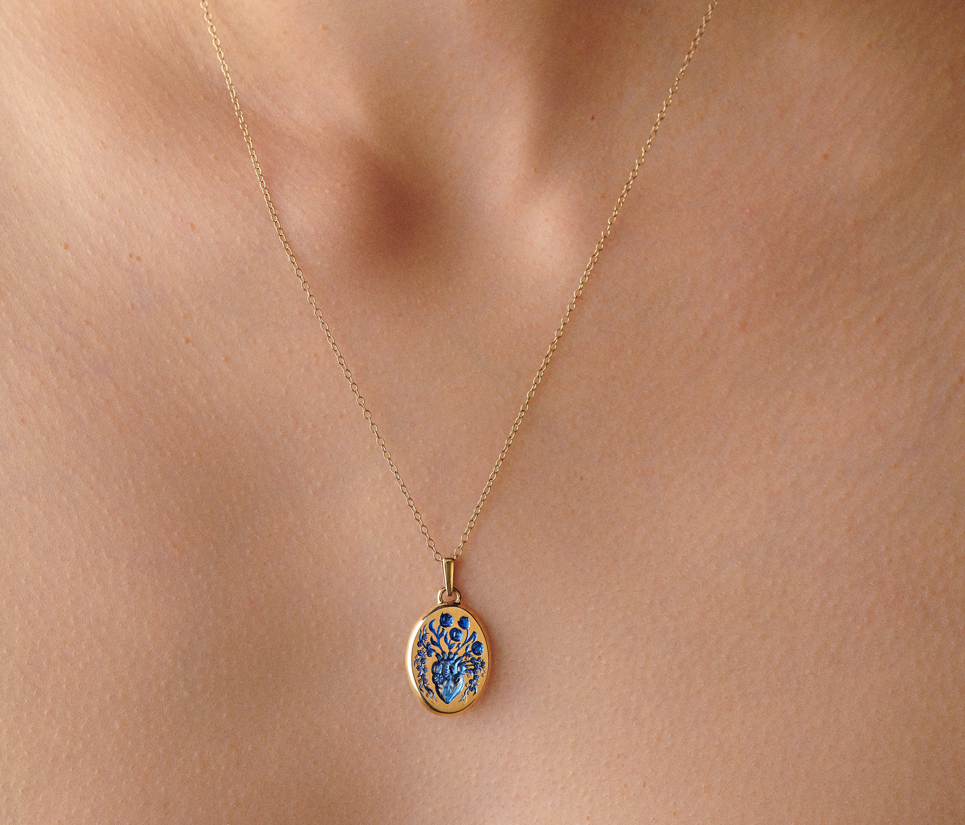 Close-up of a small gold heart pendant necklace with blue floral details worn on a person's neck