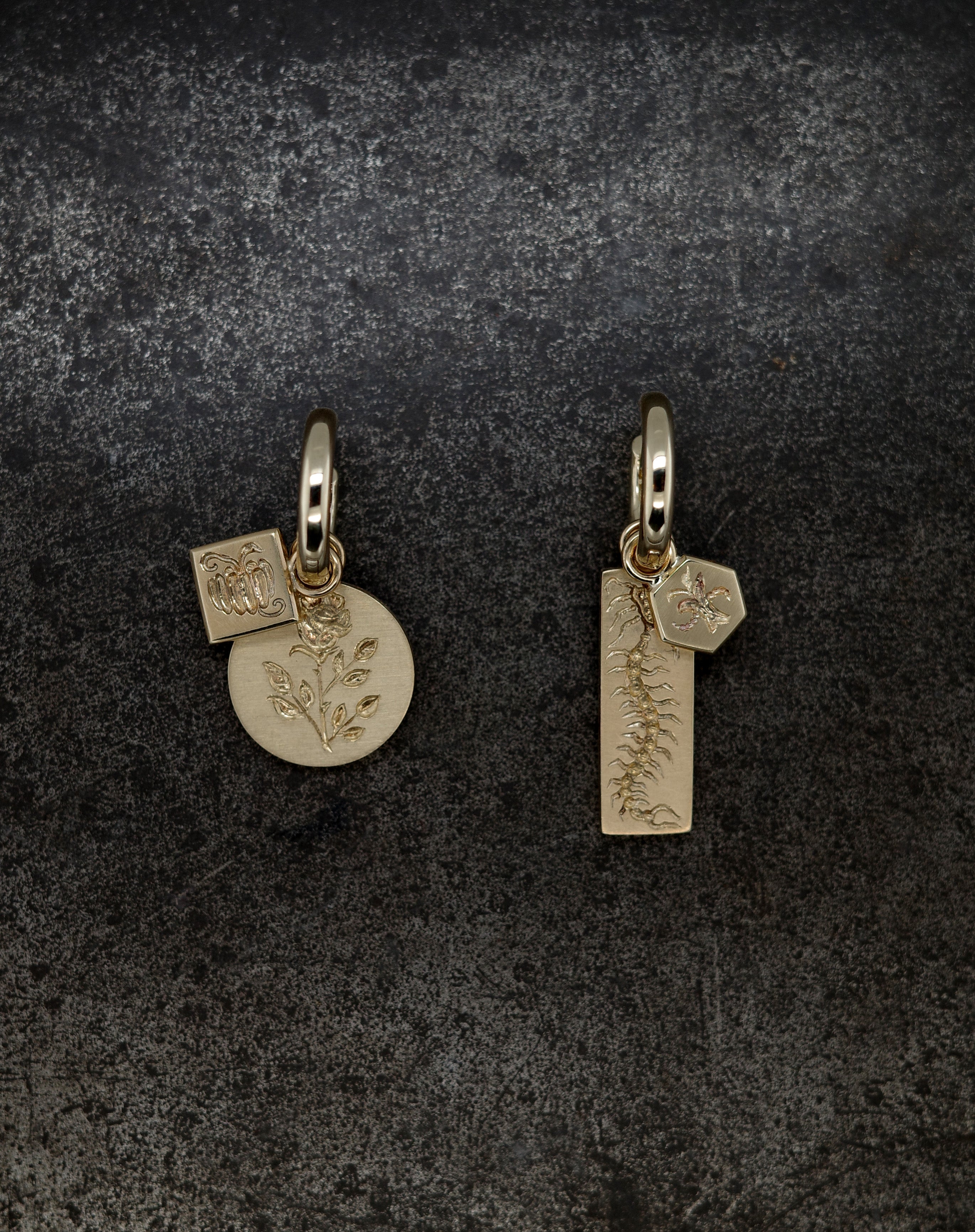 Pair of gold tiny hoop earrings with engraved botanical and centipede designs on dark textured background