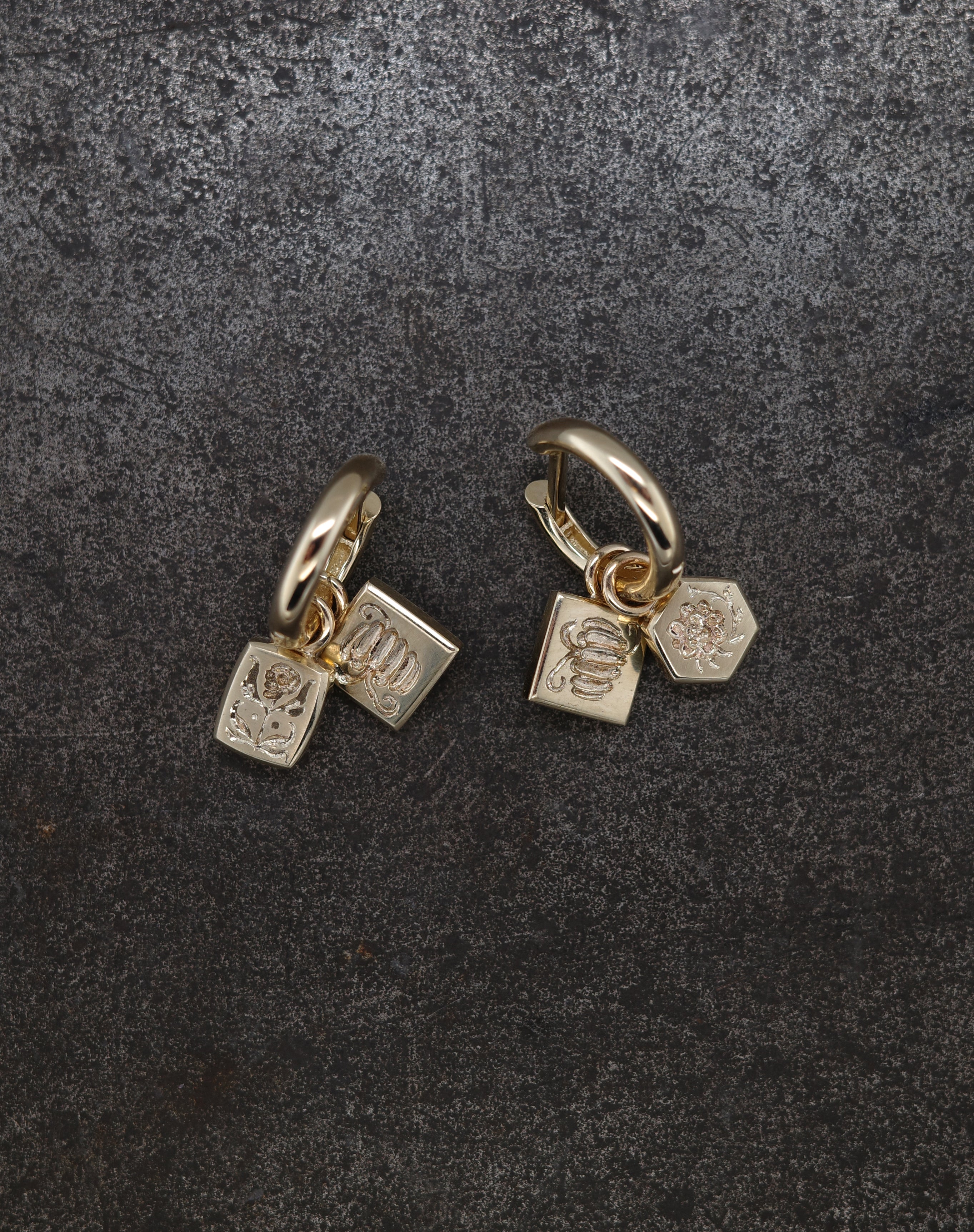 Gold-toned tiny hoop earrings with engraved pumpkin, flower, and bug charms on dark textured background
