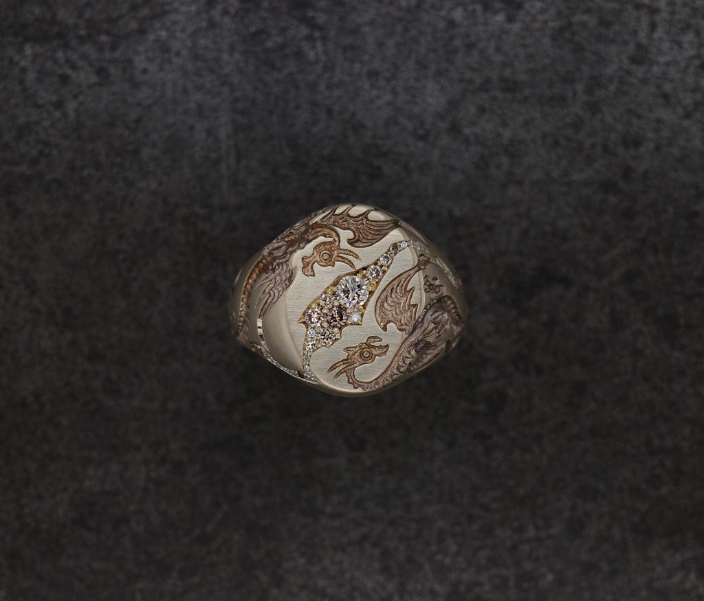 A signet ring with a cream-colored top featuring brown dragon illustrations and a central cluster of clear stones, set against a dark textured background.