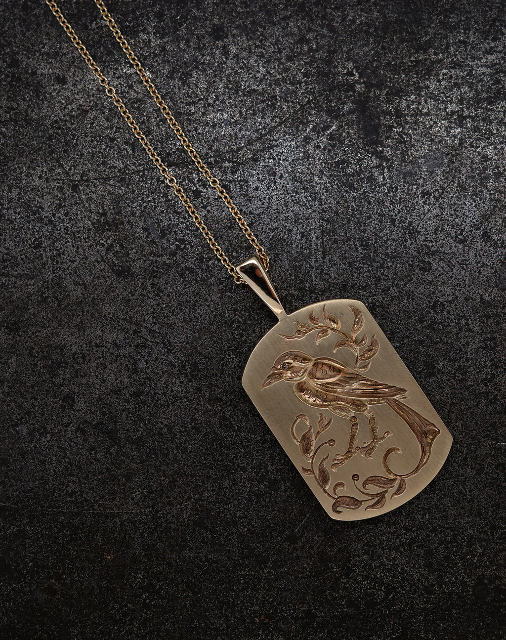 Gold bird pendant necklace with intricate floral engraving on a dark textured background