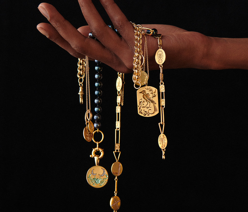 Hand holding multiple gold necklaces with intricate pendants, featuring a detailed bird pendant against a black background