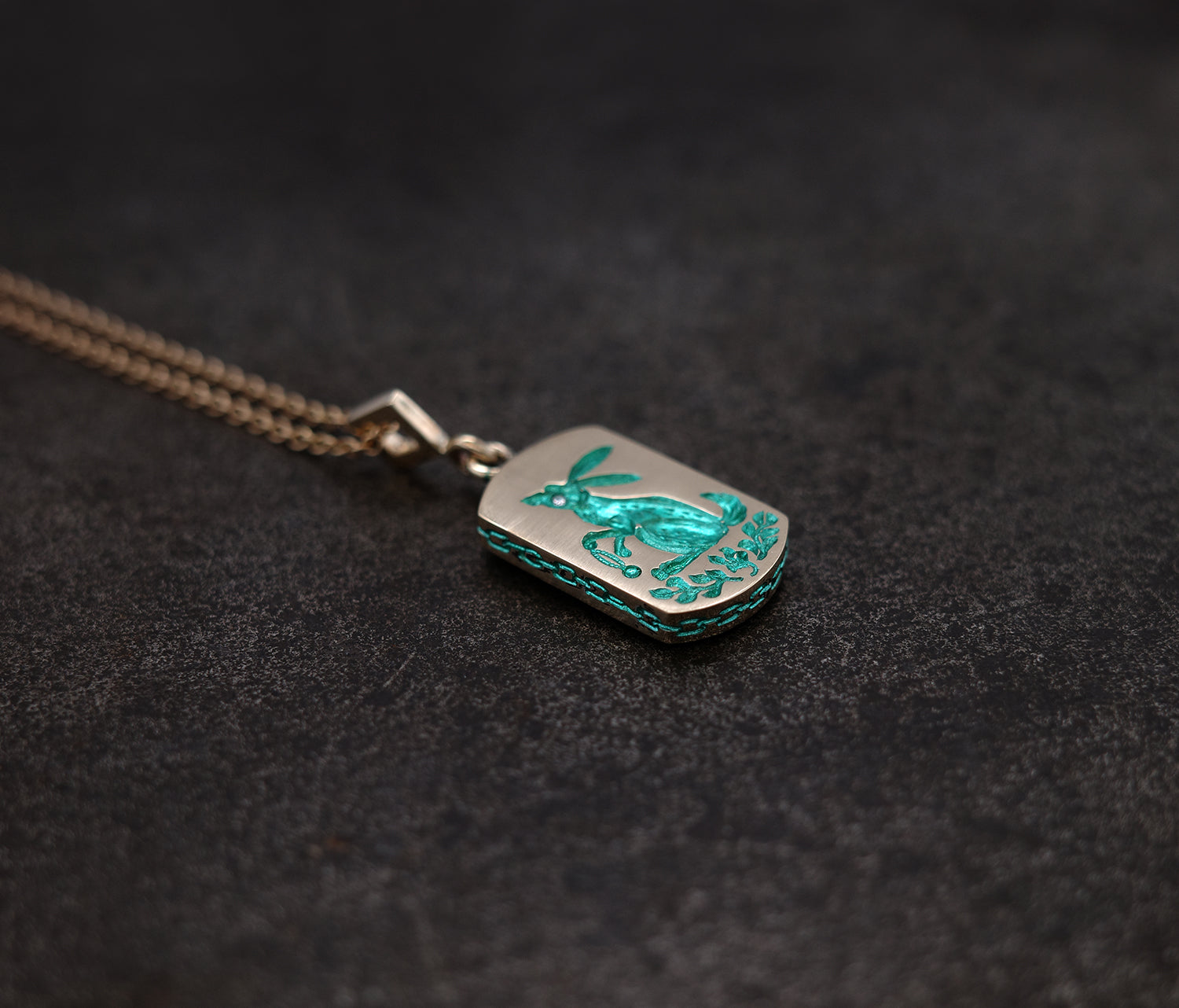 Rabbit and carrot pendant with hand-engraved design, ceramic plating, gemstone accent, and intricate chain border on dark background
