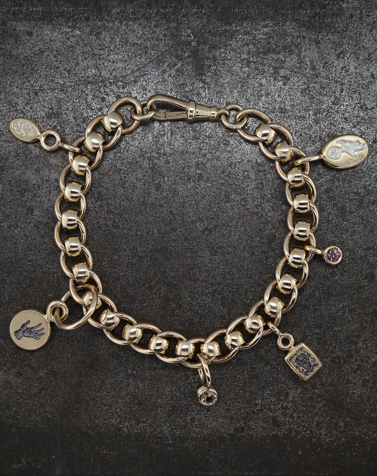 Gold chain bracelet with assorted charms including ancient talisman, hand, star, and purple gem on dark background