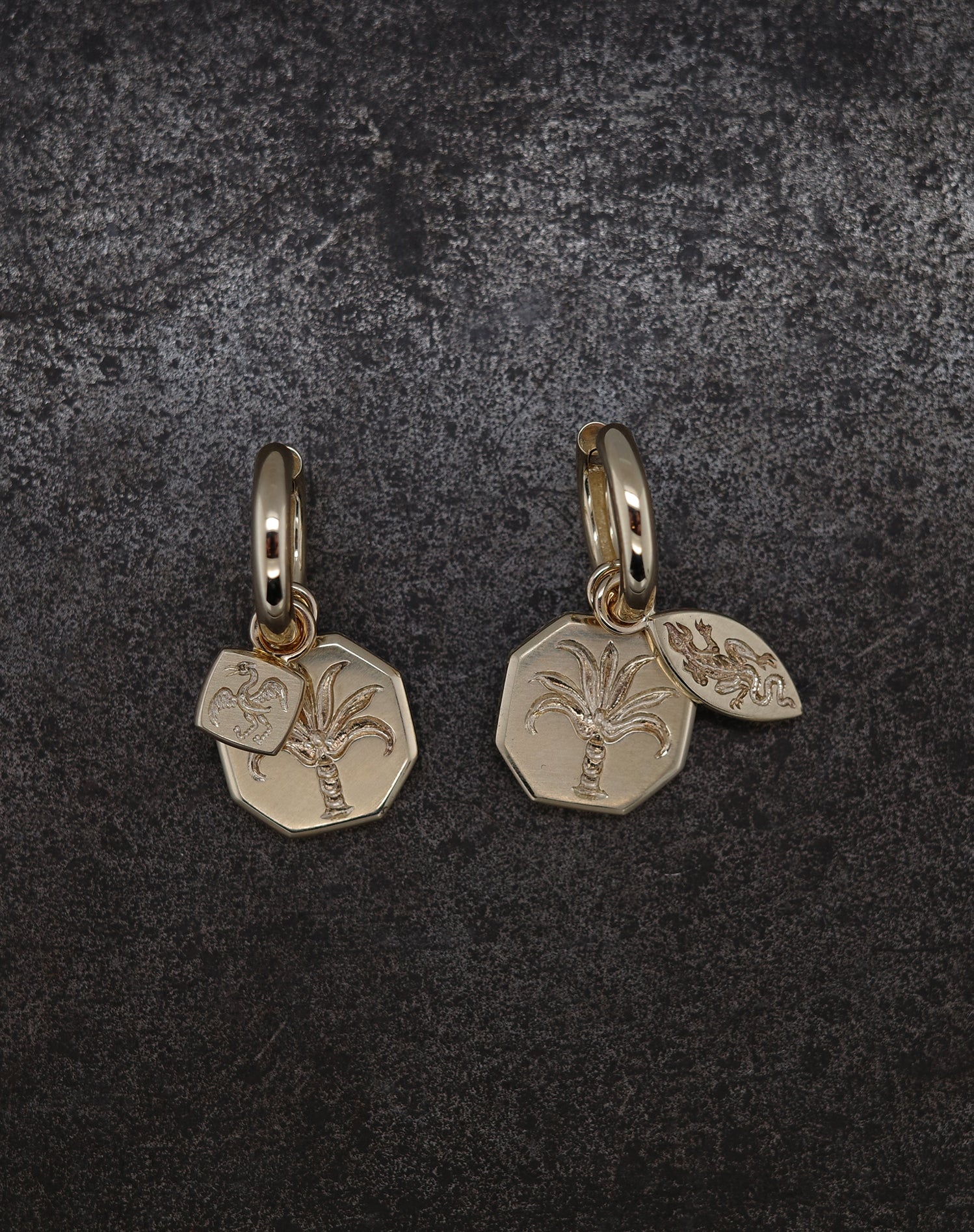 Gold hoop earrings with engraved palm tree and small charm featuring bugs and fruits design on dark background